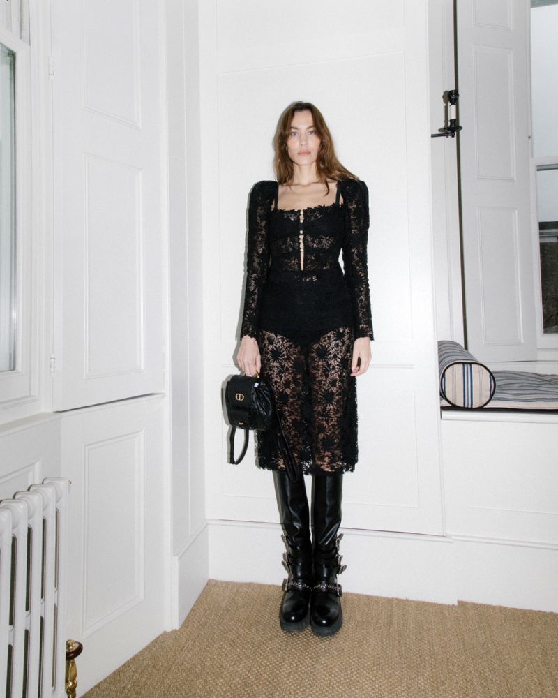 Woman posing in black lace dress with high-heeled boots and a small black bag inside an elegant white room with natural light.