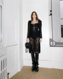 Alexa Chung wearing a black lace dress and tall boots holding a black handbag inside a room with white panelled walls.