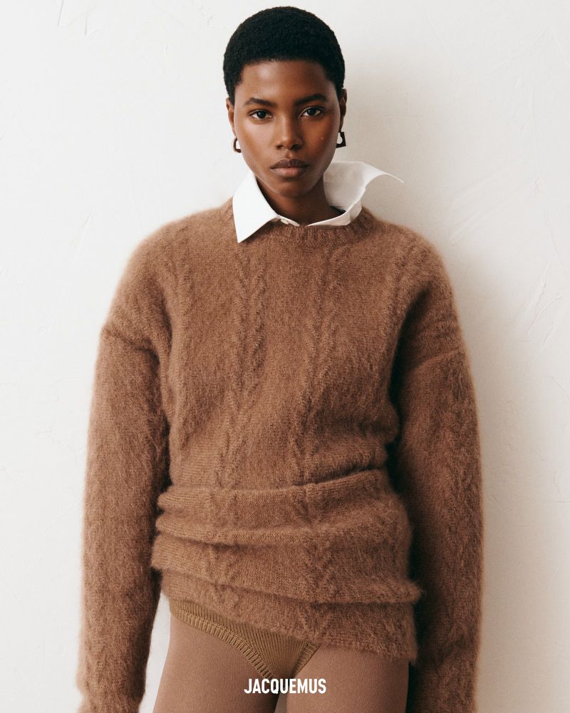 An adult female model with short dark hair poses in an indoor setting. She is wearing a brown fuzzy sweater and a white collared shirt.