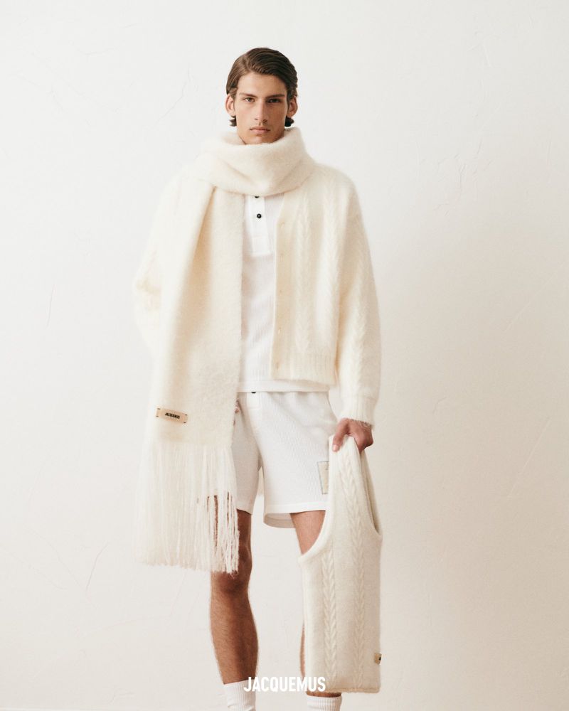 Male model in cream-colored knit cardigan, polo shirt, and scarf, holding sweater, posing against a white studio backdrop.