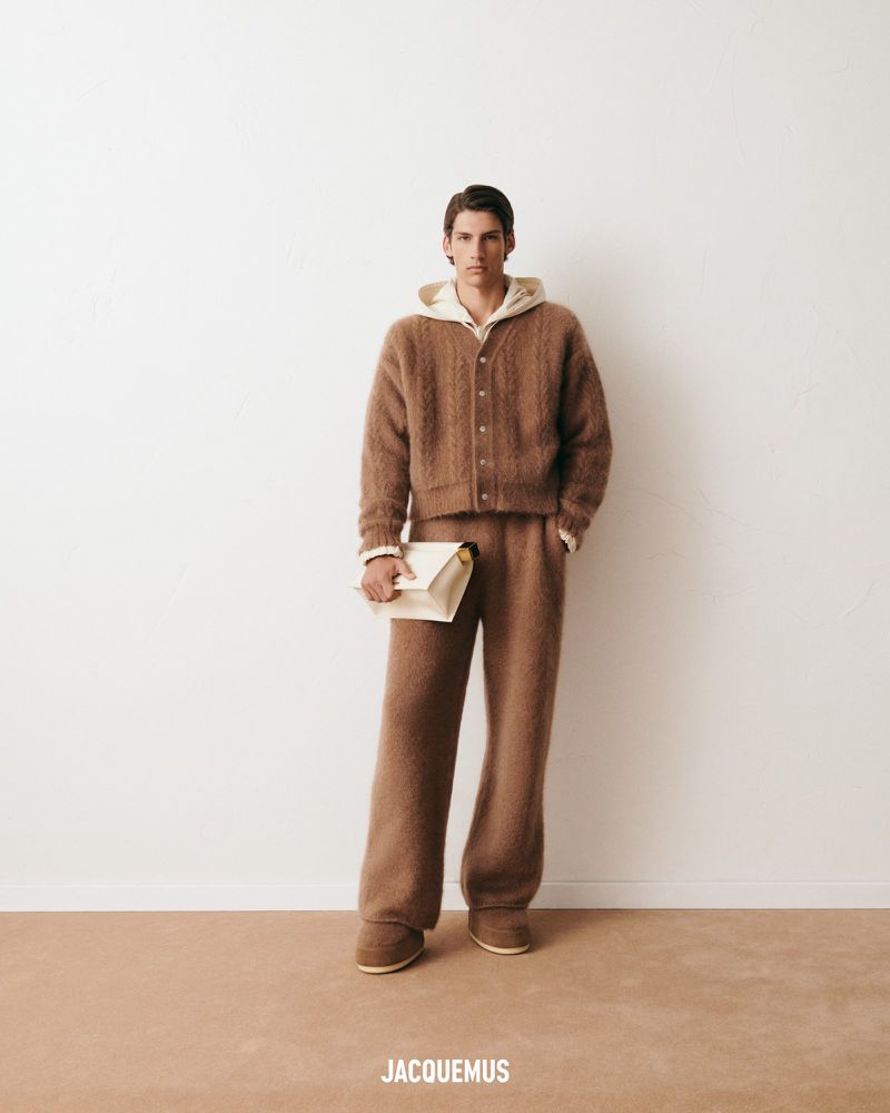 Male model wearing a brown knit cardigan, hoodie, and pants, holding an off-white clutch, against a white studio backdrop with the JACQUEMUS logo.