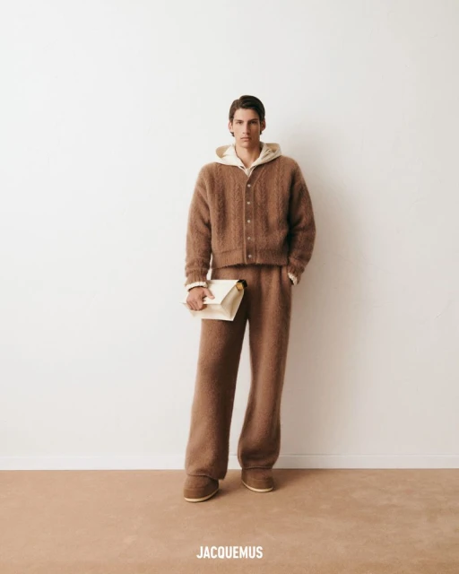 Male model wearing a brown knit cardigan, hoodie, and pants, holding an off-white clutch, against a white studio backdrop with the JACQUEMUS logo.