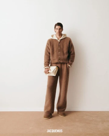 Male model wearing a brown knit cardigan, hoodie, and pants, holding an off-white clutch, against a white studio backdrop with the JACQUEMUS logo.