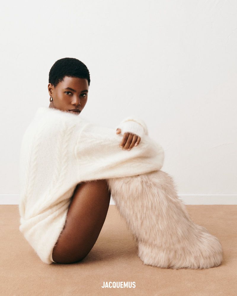 An adult woman with short dark hair poses seated, wearing a white sweater and fuzzy boots against a neutral background.