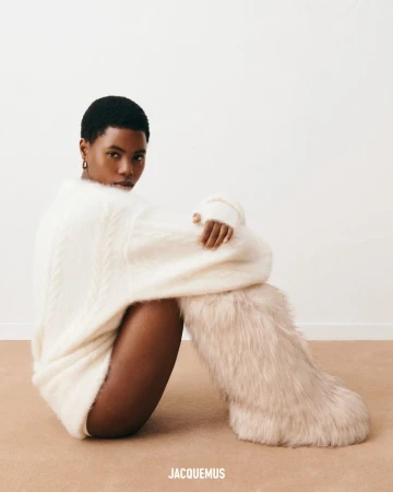 An adult woman with short dark hair poses seated, wearing a white sweater and fuzzy boots against a neutral background.
