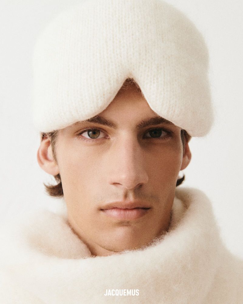 Close-up studio shot of a young adult man wearing a white knit balaclava, a white knit sweater, and featuring the brand 'Jacquemus'.