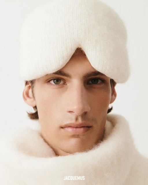 Close-up studio shot of a young adult man wearing a white knit balaclava, a white knit sweater, and featuring the brand 'Jacquemus'.