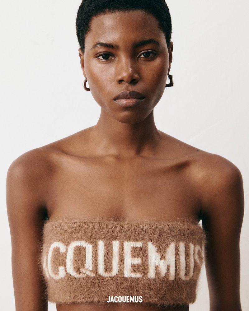 Close-up portrait of a Black female model wearing a brown knit bandeau top with white lettering against a plain white background.