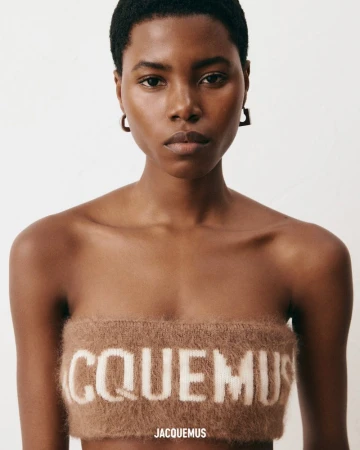 Close-up portrait of a Black female model wearing a brown knit bandeau top with white lettering against a plain white background.
