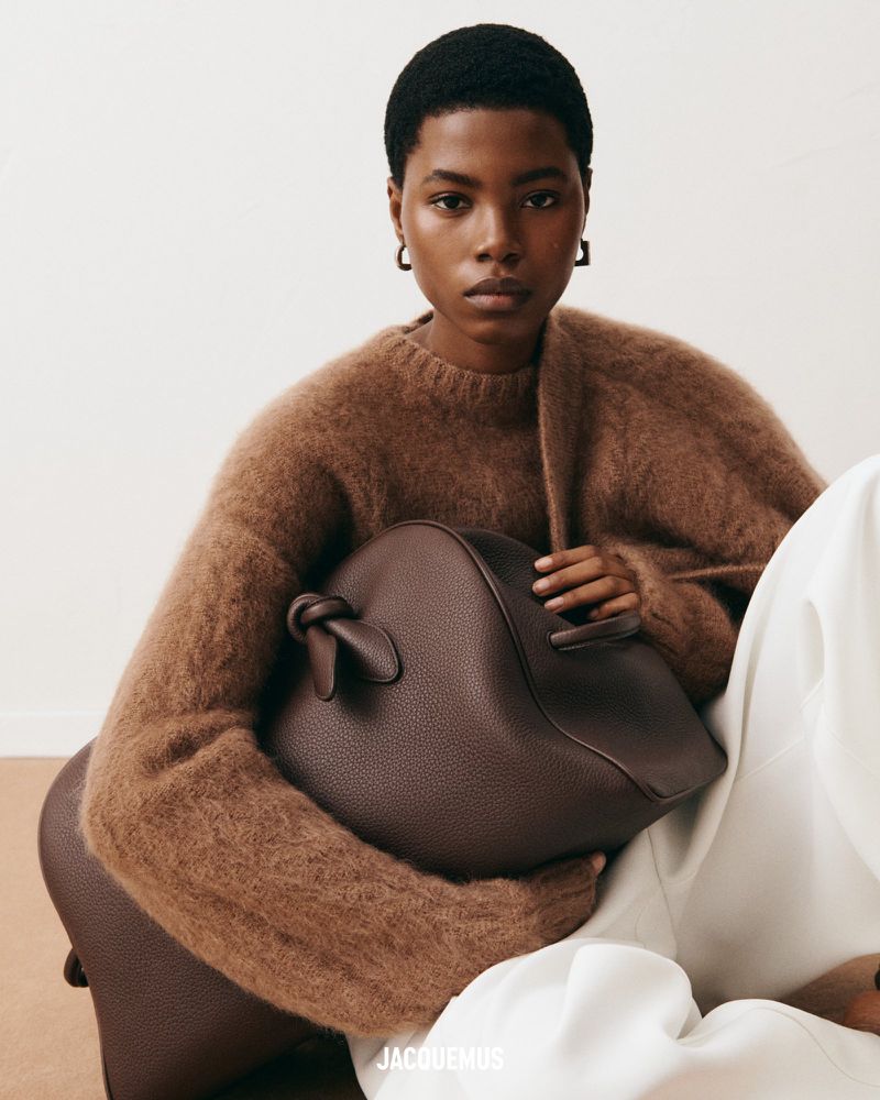 Fashion image of a Black woman in a brown sweater holding a brown purse on a light background, with the brand logo visible.