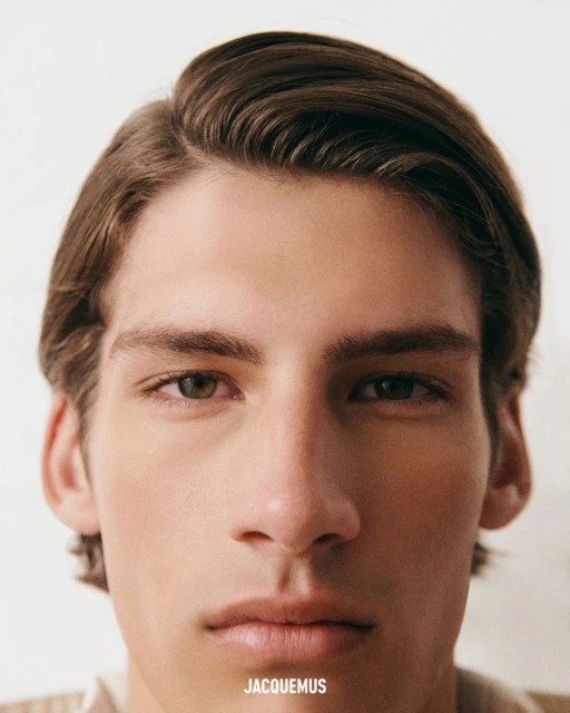 Close-up studio portrait of a young adult male with medium-length brown hair, wearing a light-colored shirt and posing against a white backdrop.