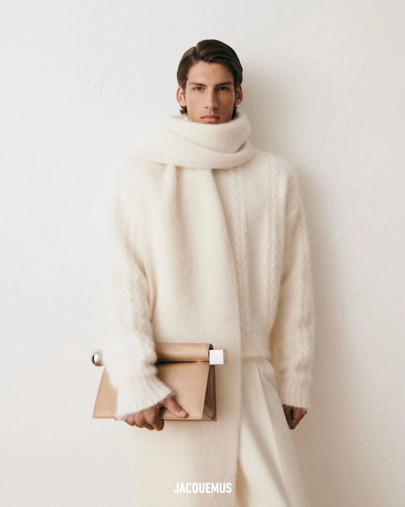 Model wearing a thick white knit sweater and matching scarf while holding a tan leather clutch against a plain wall.