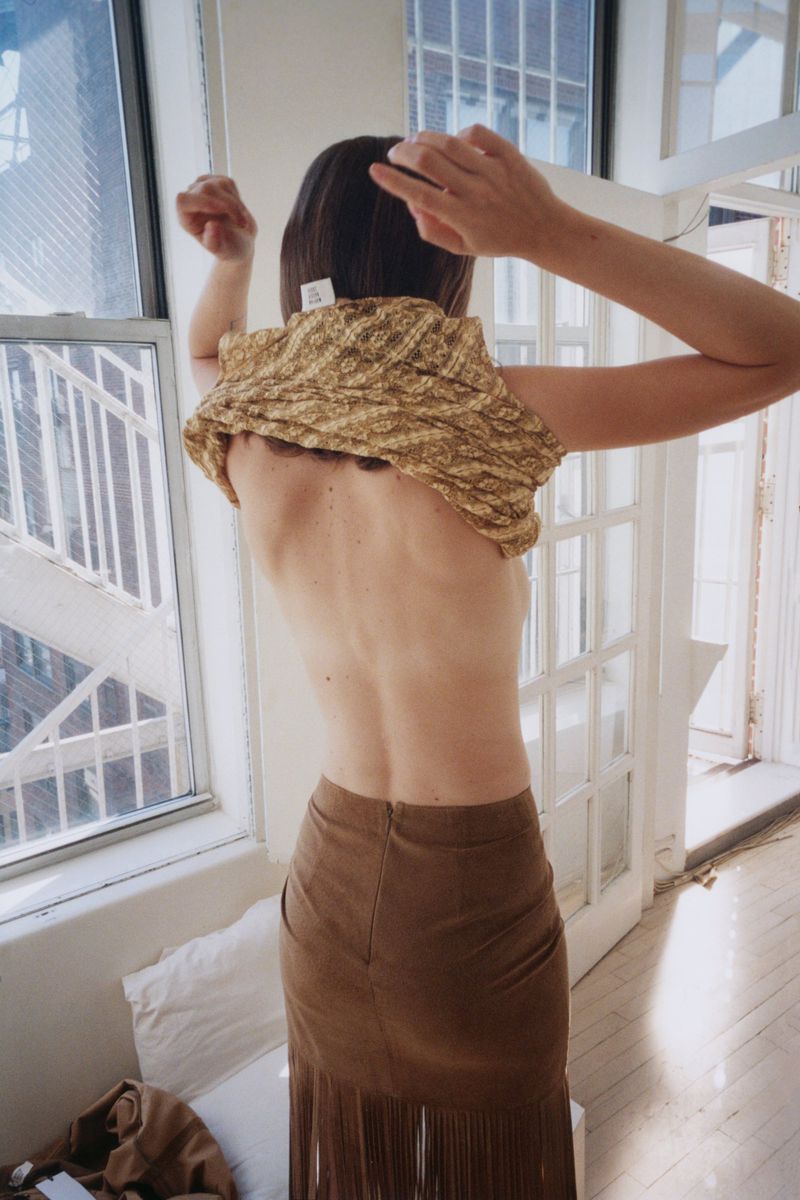 Back view of an adult woman changing her top in an indoor setting with light brown hair, a brown skirt with fringe, and natural lighting.