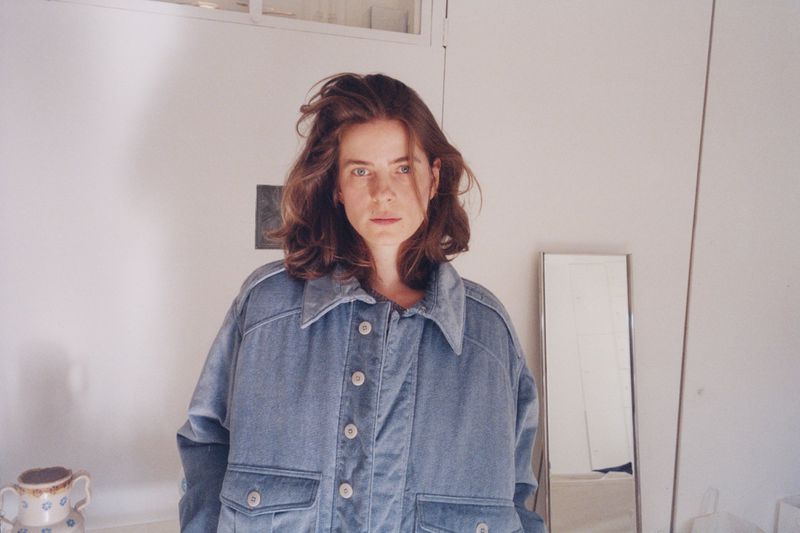 Woman wearing a blue velvet button-down jacket standing in a bright minimalist interior with a mirror