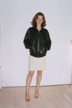 A woman in a black leather jacket and neutral skirt poses in a full shot against a stark white background with minimalist styling.