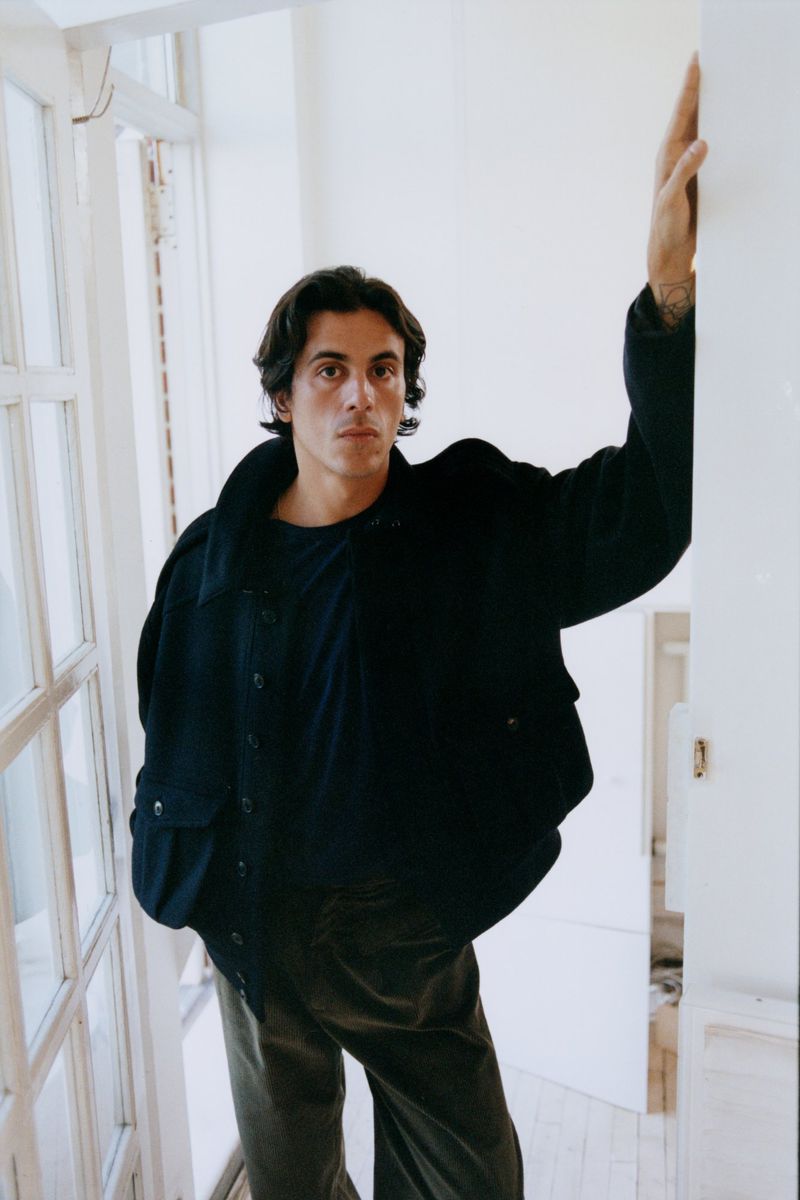 Adult male posing in a dark jacket and pants, stands next to a white door frame, indoor lighting, studio shot.