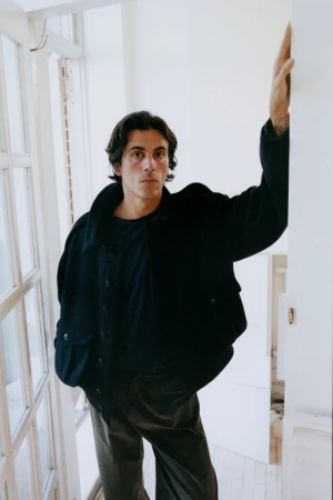 Adult male posing in a dark jacket and pants, stands next to a white door frame, indoor lighting, studio shot.