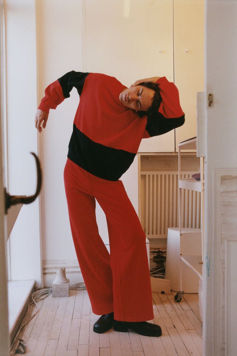 Full-length shot of a person in a red and black sweater and red pants striking a pose in an indoor environment with a white wall backdrop.