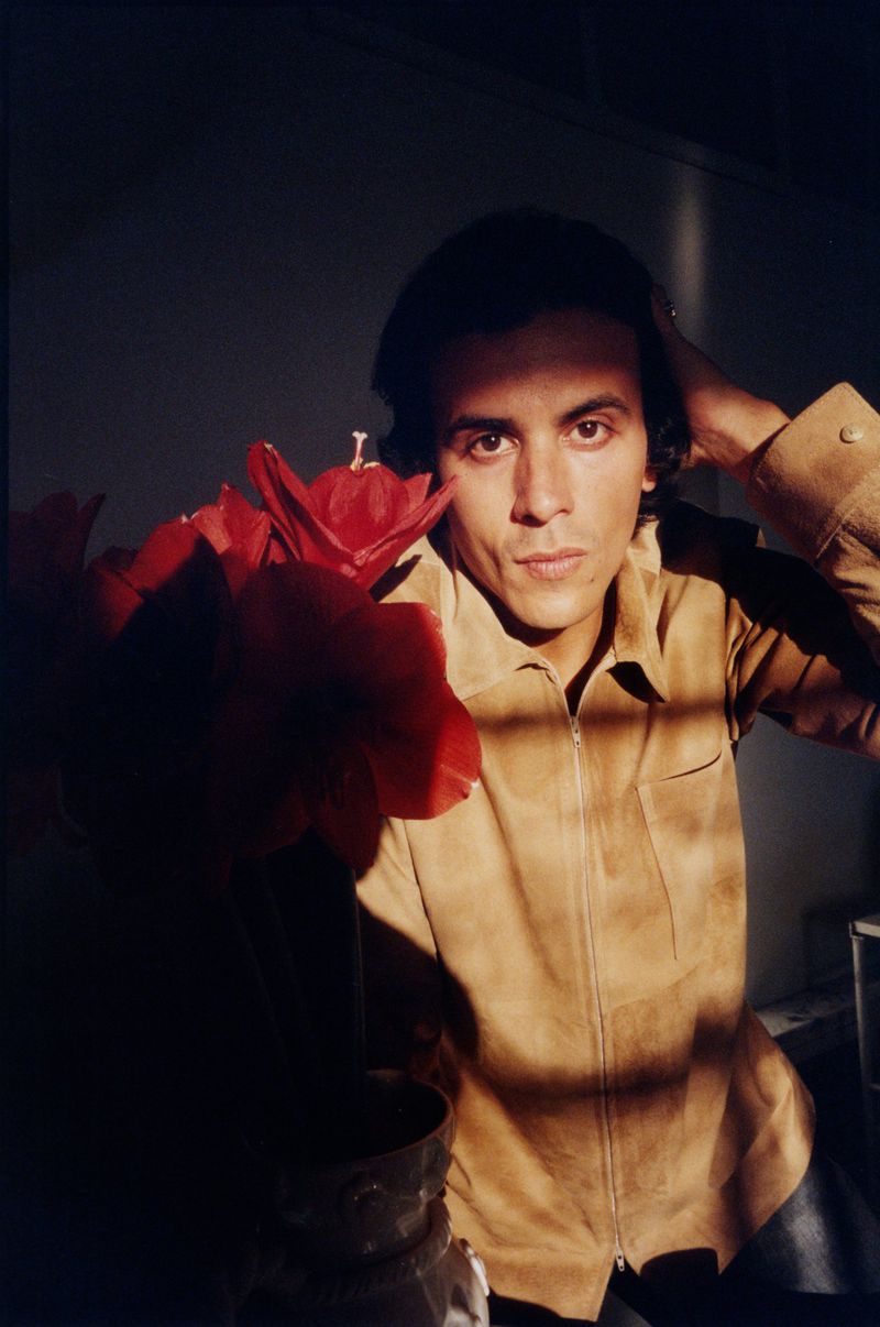 Man in a tan suede shirt posing beside a vase of red flowers with dramatic window light casting shadows across his face and jacket.