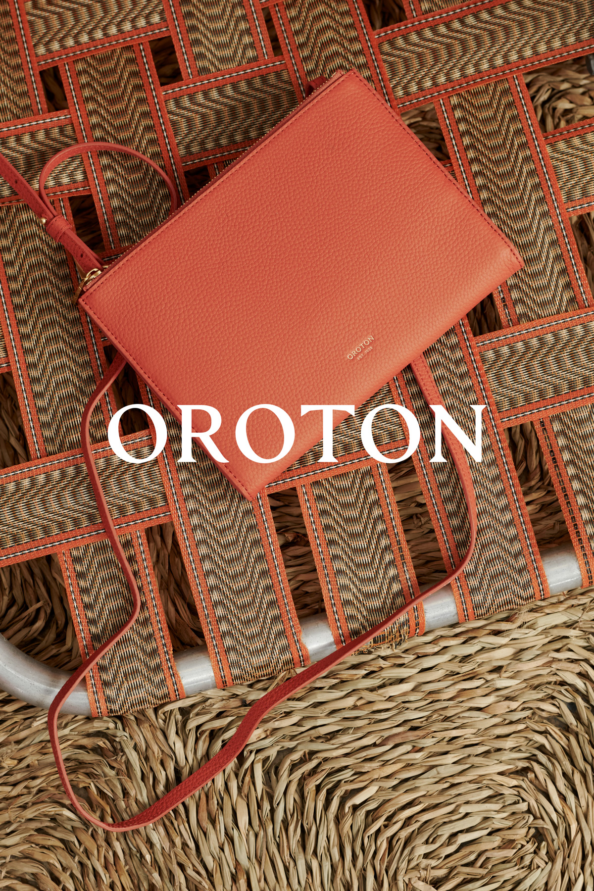Elevated view of a coral-colored Oroton handbag with a shoulder strap placed diagonally on a woven surface. Brand name is in white.