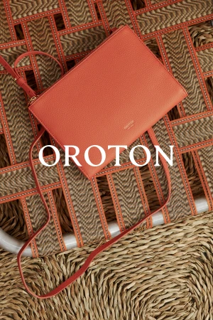 Elevated view of a coral-colored Oroton handbag with a shoulder strap placed diagonally on a woven surface. Brand name is in white.