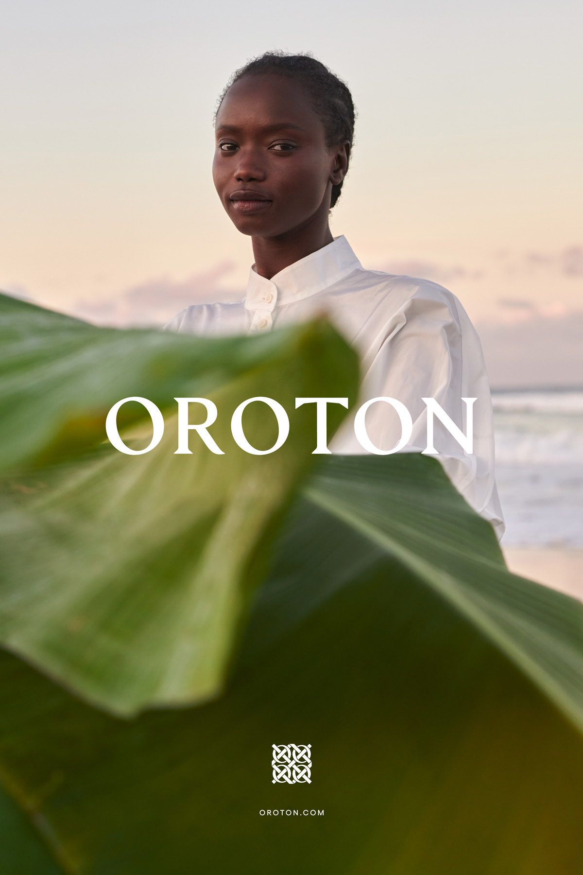 Portrait of a black woman wearing a white shirt behind large green leaves against a beach and sunset sky backdrop.