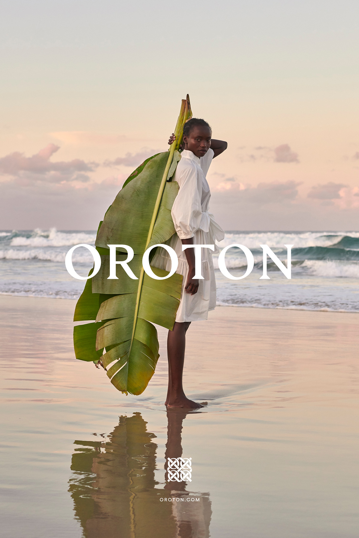 Fashion shoot on a beach featuring a Black woman in a white dress holding a large banana leaf against a sunset seascape.