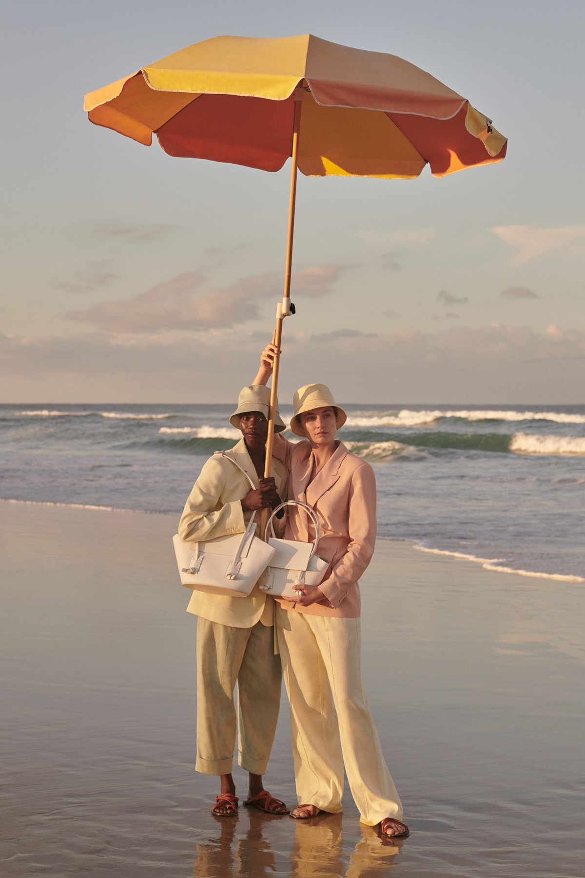 Two models posing on a beach under a large umbrella. They both wear suits, hats, and sandals, and each holds a light-colored handbag.