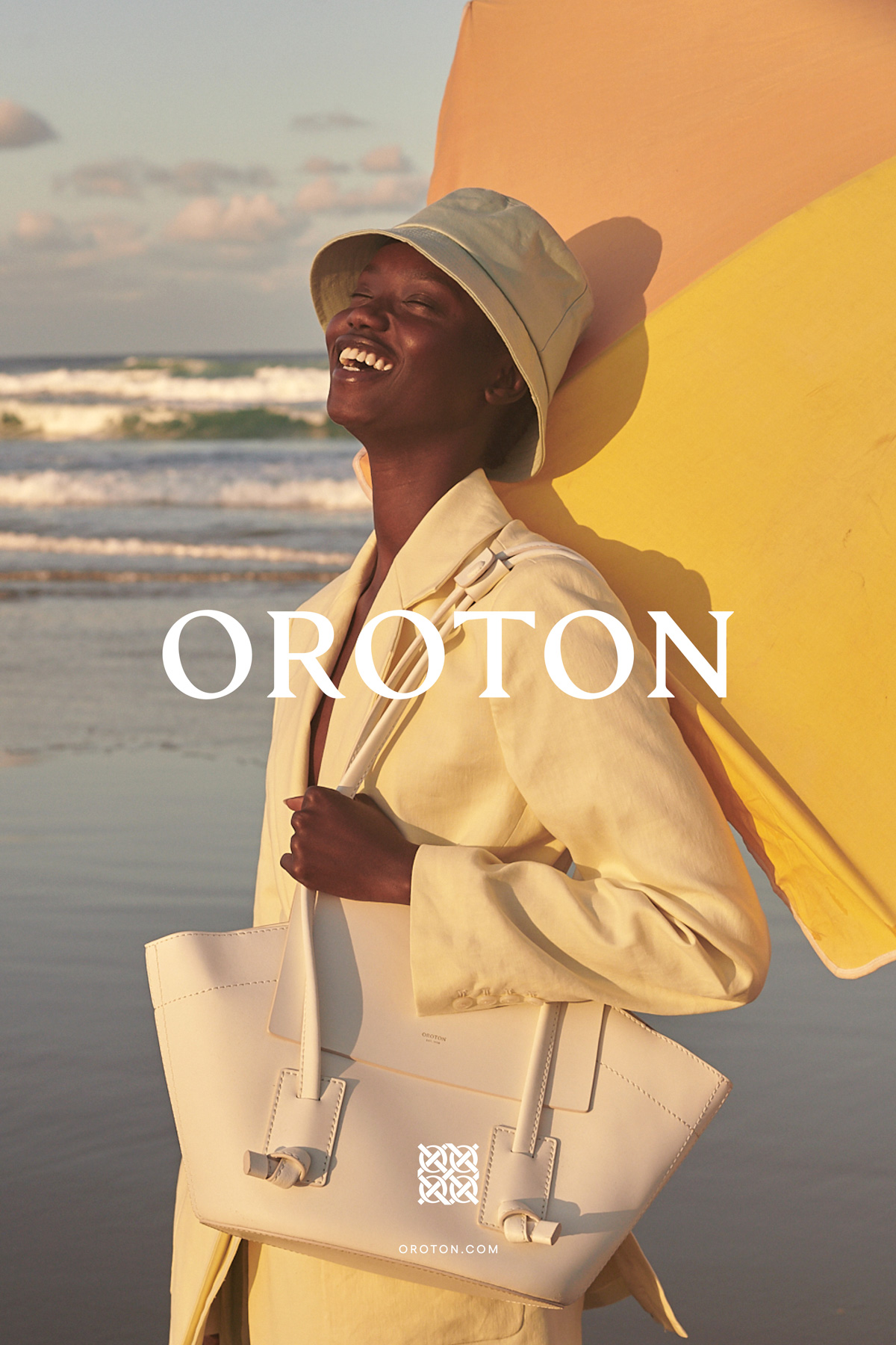 Smiling woman in bucket hat and jacket on beach with designer handbag and colorful backdrop for fashion brand Oroton advertisement.