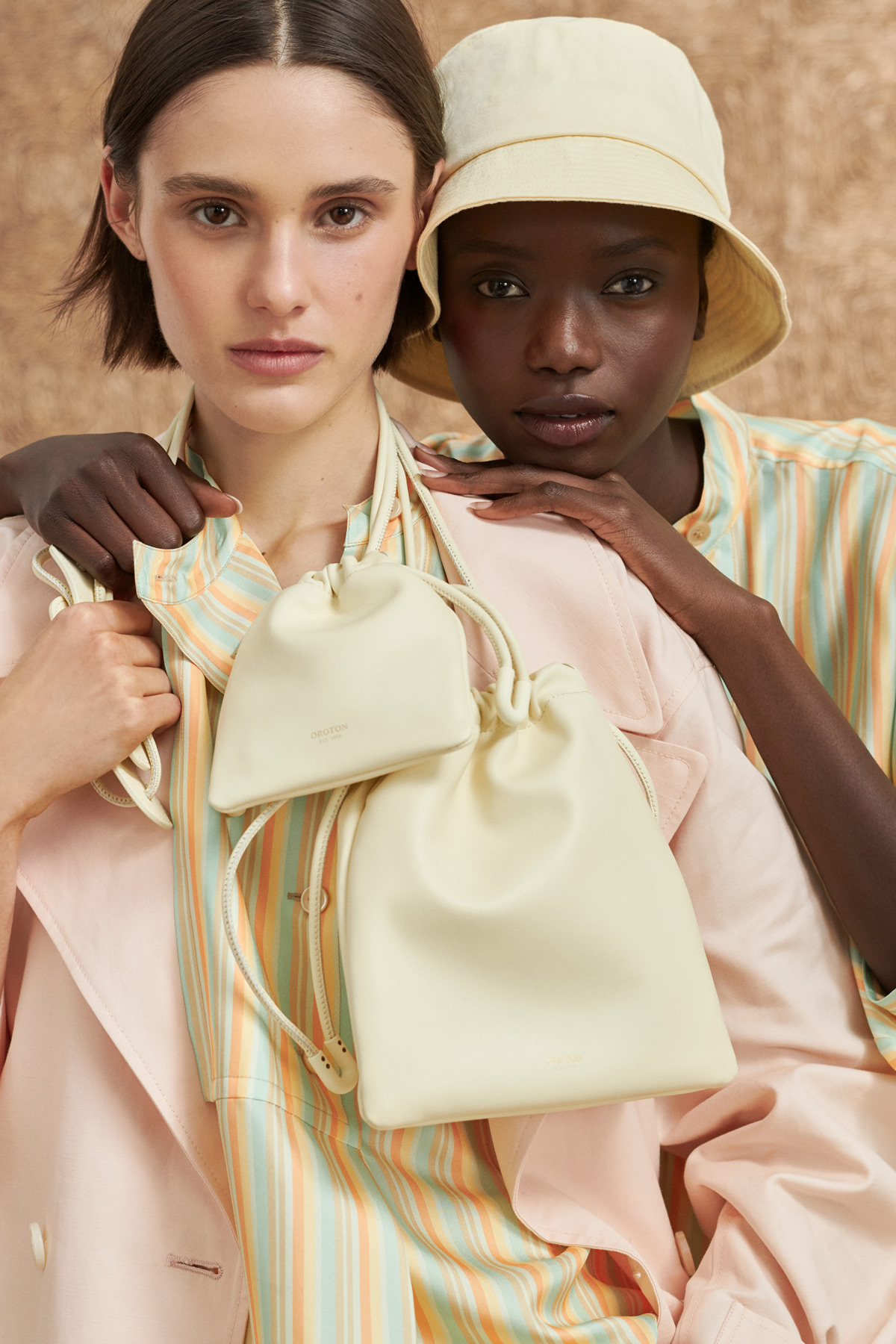 Two models pose with ivory-colored leather bucket bags in a close-up studio shot, wearing pastel striped shirts and a bucket hat.