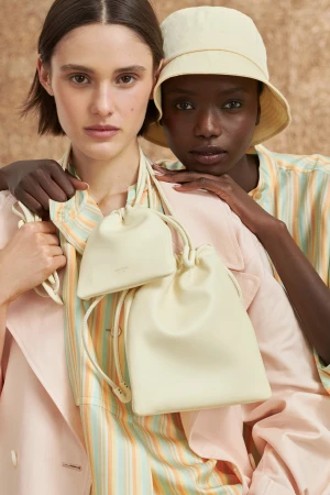 Two models pose with ivory-colored leather bucket bags in a close-up studio shot, wearing pastel striped shirts and a bucket hat.