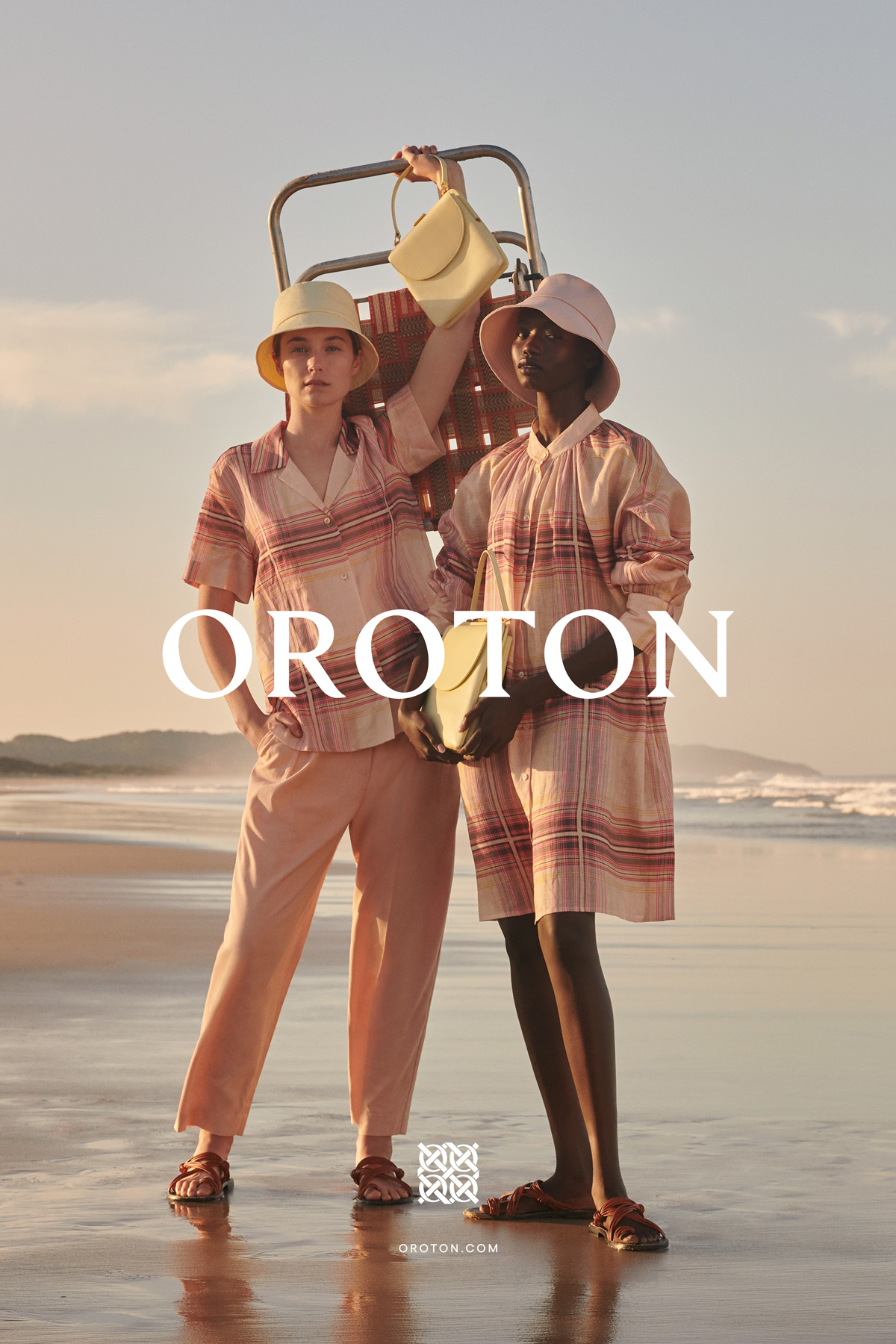 Two women pose on a beach in matching striped outfits, holding purses, with a beach chair and the Oroton logo visible.
