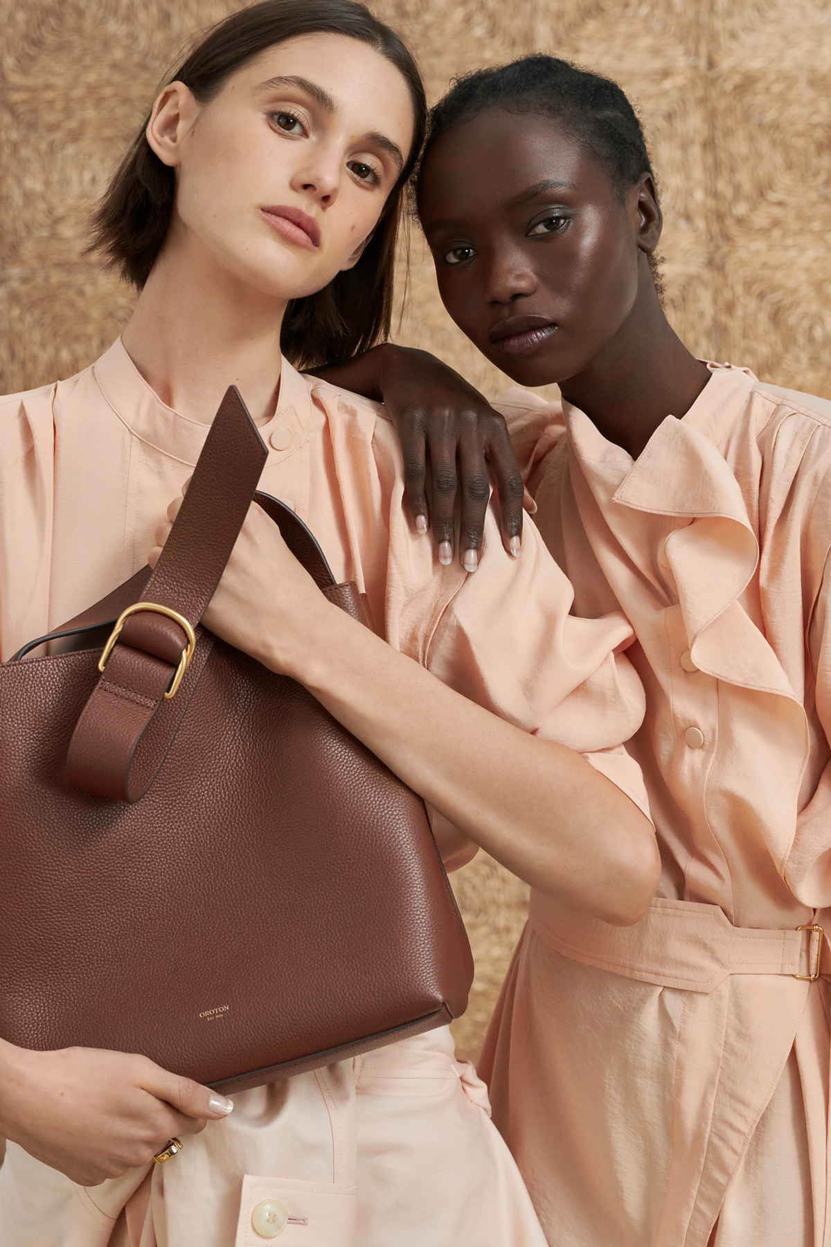Two models pose with a brown leather tote bag in a studio, showcasing fashion and accessories with a focus on texture and color coordination.