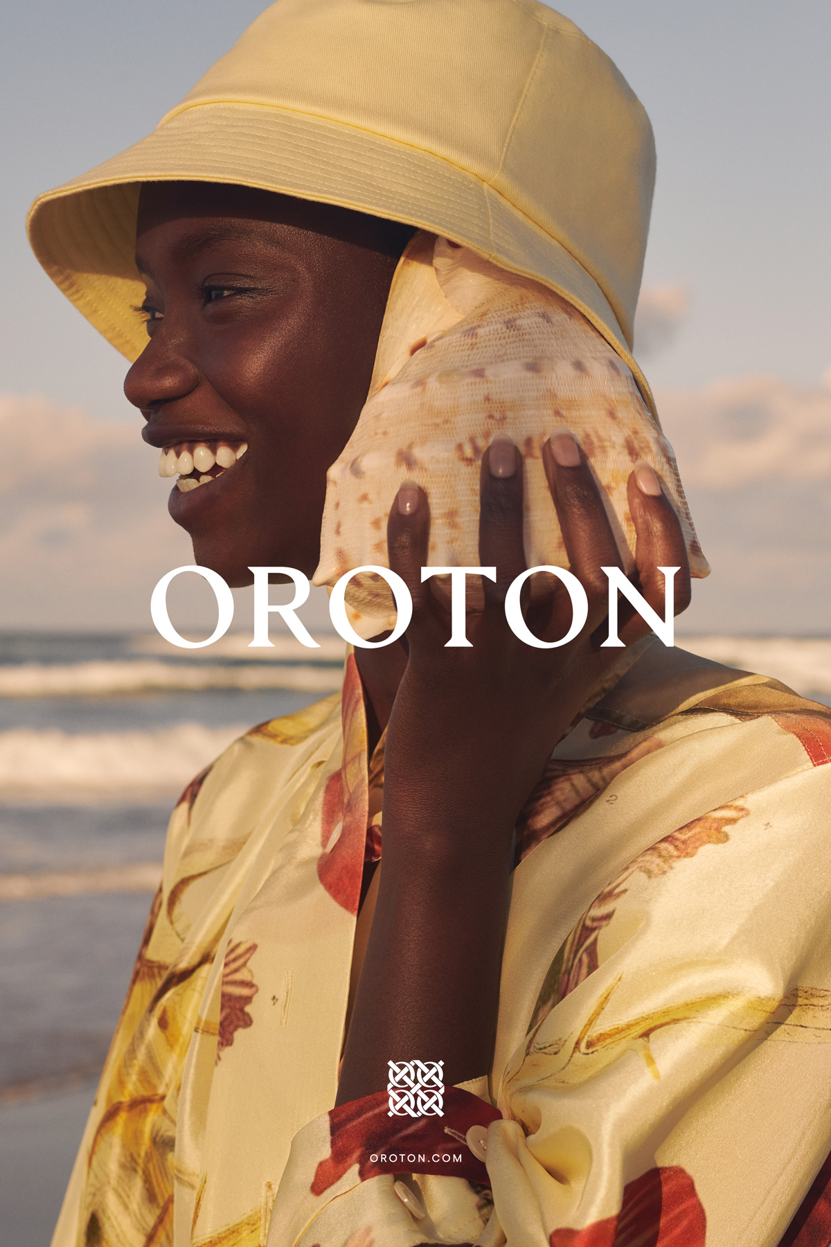 Smiling Black model in yellow hat holding seashell to her ear on beach. She wears a floral blouse by Oroton. Sea and sky background.