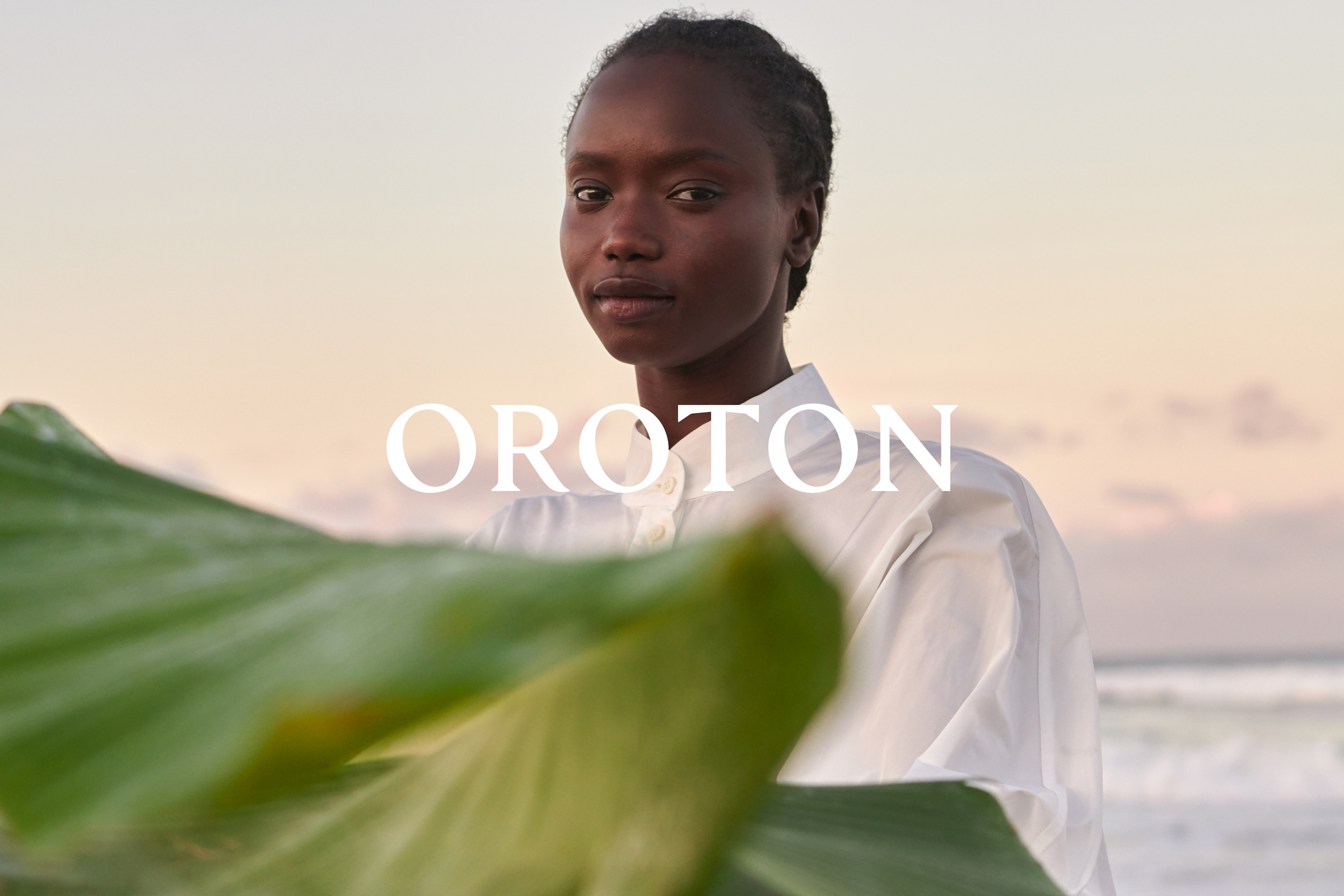 Portrait of an African woman in white shirt with a large green leaf in the foreground, against a blurred background of sky and sea.