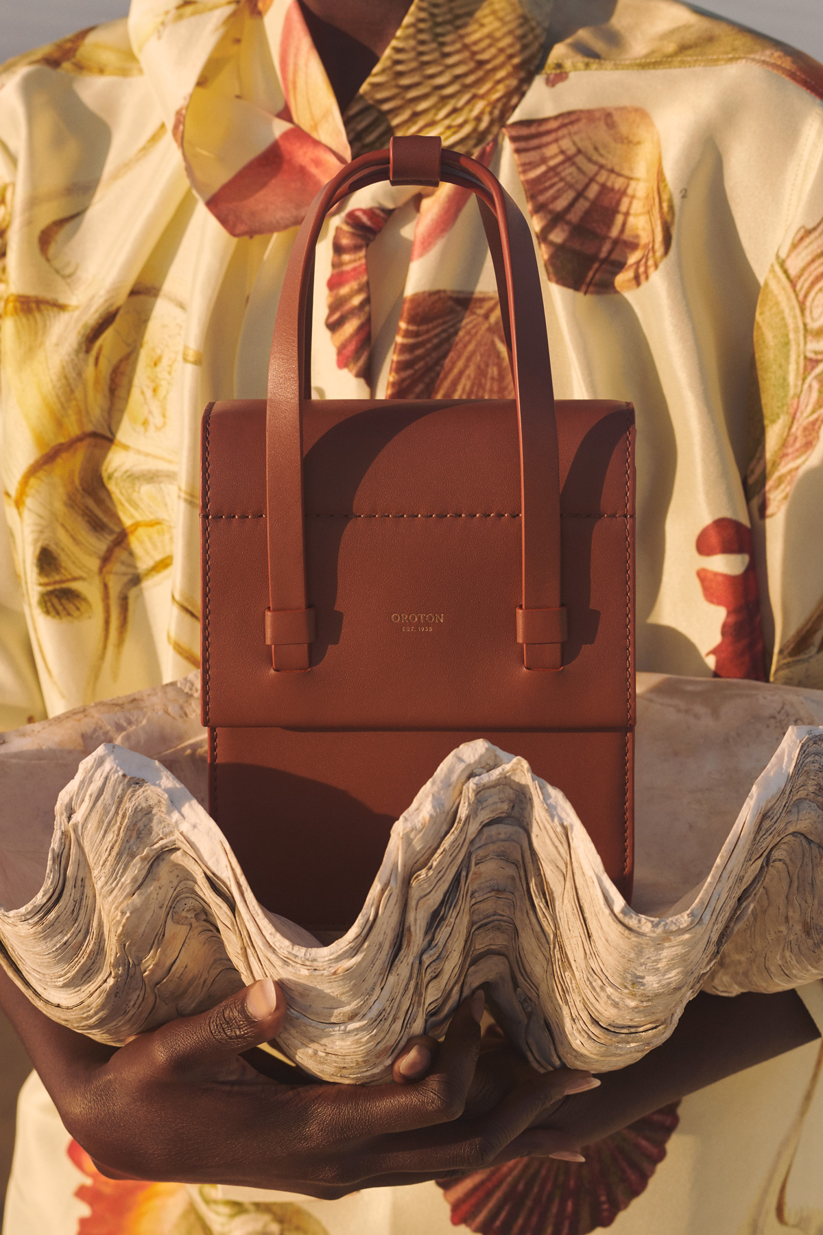 A sophisticated brown leather purse with top handles is presented against a background of a seashell-patterned shirt and a large clam shell.