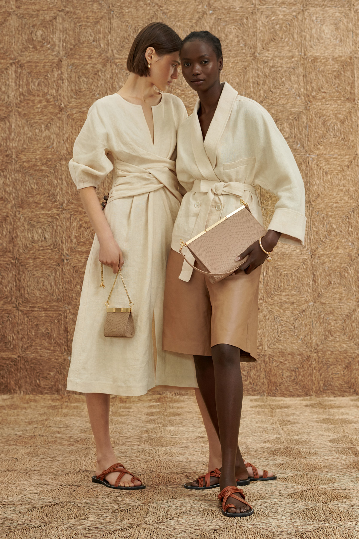 Two women pose in neutral-toned dresses and sandals, each holding a handbag, against a textured straw-like backdrop, creating a fashion editorial.