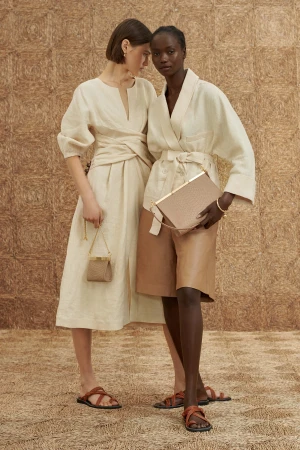 Two women pose in neutral-toned dresses and sandals, each holding a handbag, against a textured straw-like backdrop, creating a fashion editorial.