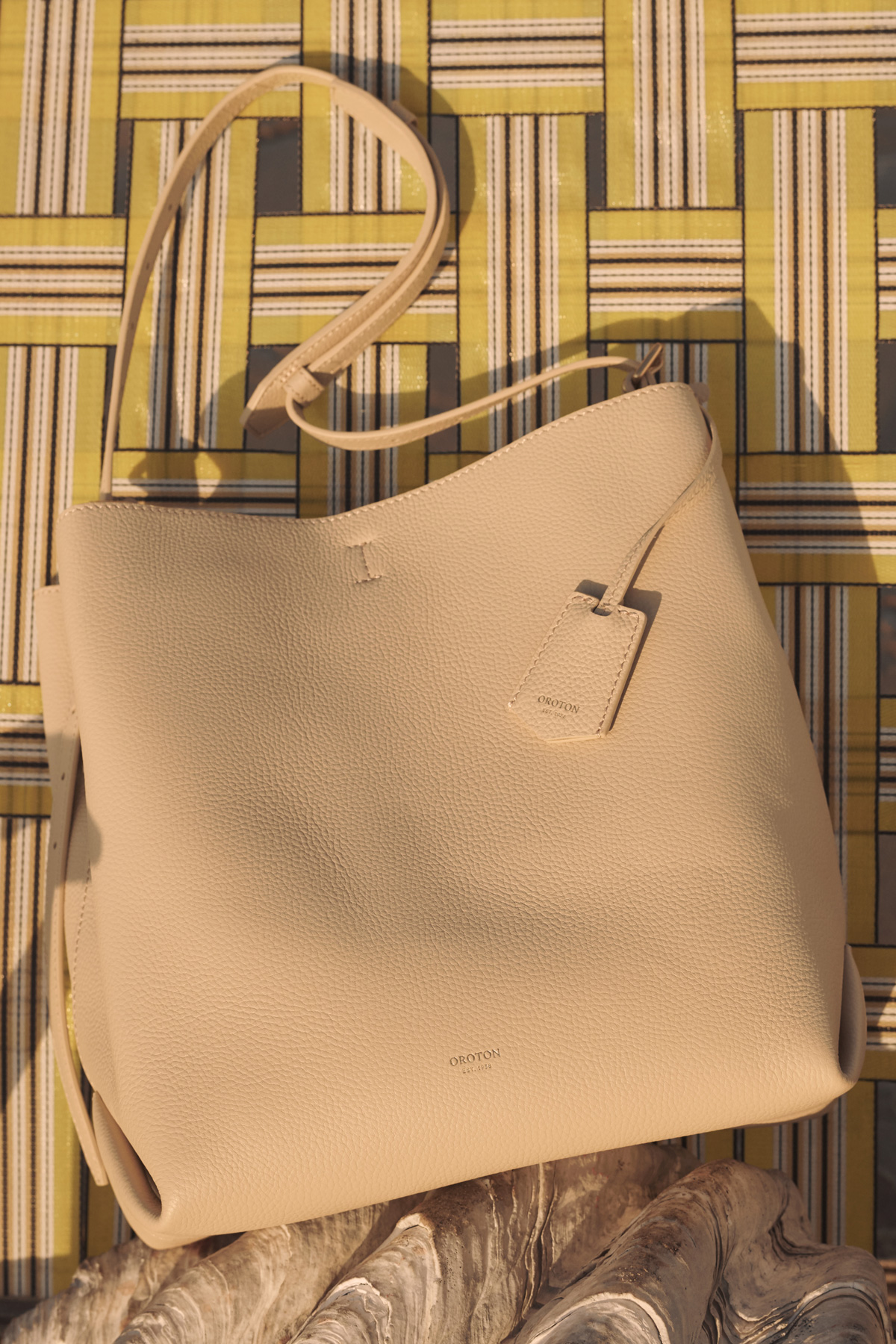 Beige faux leather hobo handbag with shoulder strap and tag in a studio setting against a patterned background and seashell.