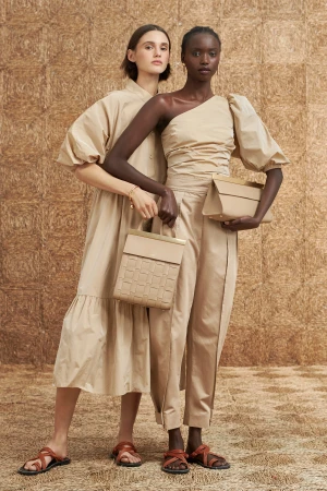 Two women in neutral dresses and sandals holding matching handbags against a textured background in a fashion editorial shot.