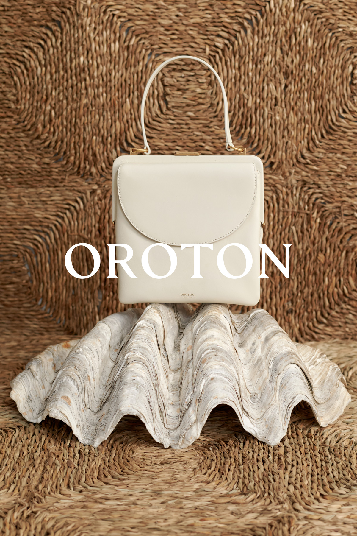 Cream-colored Oroton handbag with top handle and clasp closure, presented on a large shell against a woven straw background.