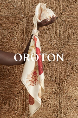 Dark-skinned hand holds a shell tied with a patterned scarf against a woven wall; Oroton text overlay at center.