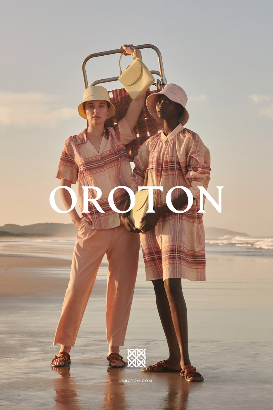 Two female models posing on a sandy beach at sunset wearing matching plaid resort wear, bucket hats, and holding leather handbags.