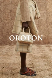Model holding two matching beige plaid handbags wearing a plaid skirt and brown sandals against textured backdrop