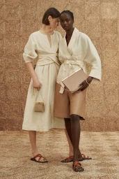 Two female models wearing cream linen outfits and brown leather sandals posing against a textured woven backdrop for Oroton Summer.