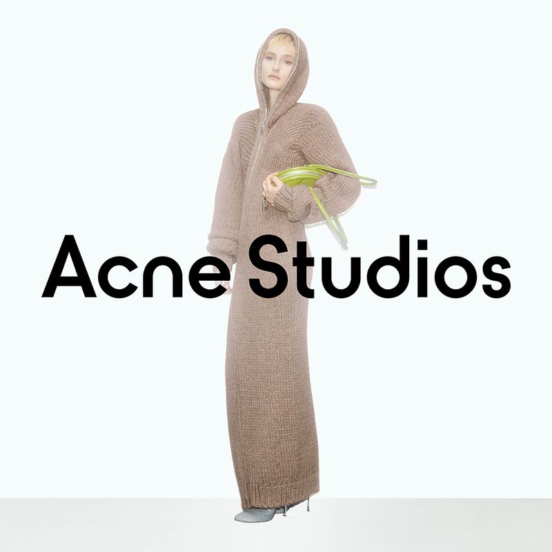 Adult female fashion model poses in a long hooded brown knit dress and gray flats, holding green palm fronds against a white backdrop.