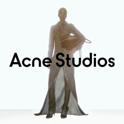 Model wearing a long denim coat holding a large dark handbag against a seamless white background for Acne Studios