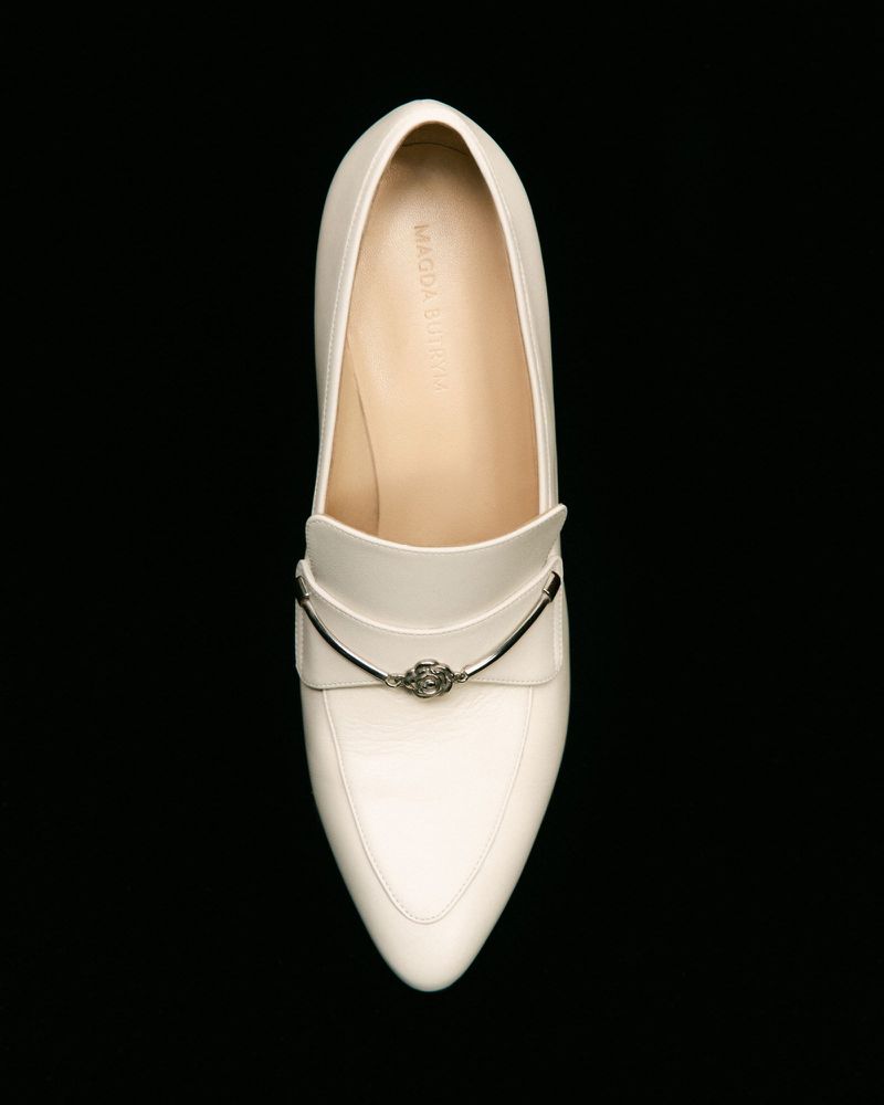 Elevated view of a single off-white leather loafer with a silver metal accent on a solid black backdrop in a studio setting.