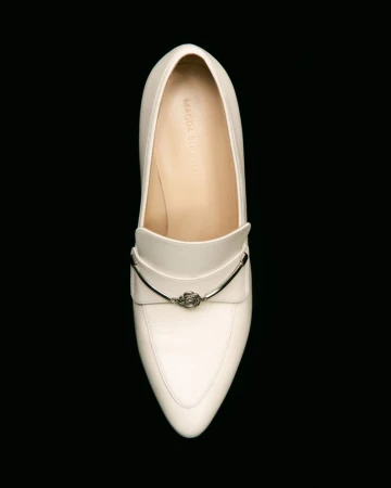Elevated view of a single off-white leather loafer with a silver metal accent on a solid black backdrop in a studio setting.