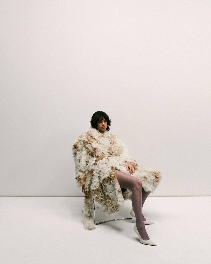A young adult woman with dark hair sits on a metal chair wearing a fuzzy, oversized coat and matching heels in a brightly lit studio.